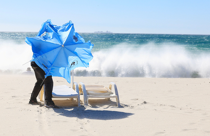 Мужчина пытается открыть зонтик во время сильных порывов ветра «Кейп-Доктор», дующего на пляже Кэмпс-Бэй в Кейптауне.